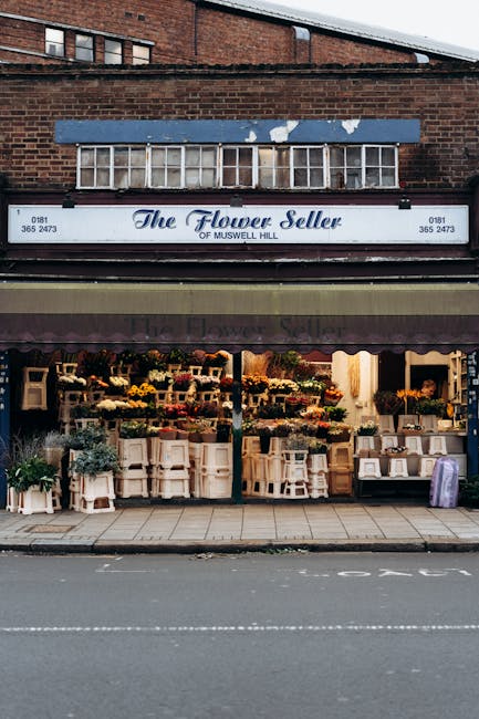 The storefront of The Flower Seller of Muswell Hill featuring a wide display window and signage, situated on a brick building. Outside, a variety of floral arrangements are arranged on white wooden st