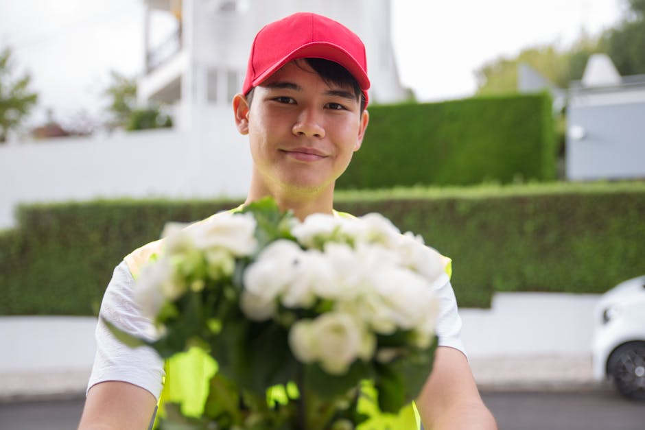 A delivery person wearing a red cap and a high-visibility vest is holding a large bouquet of white flowers with green foliage, extending it towards the camera with a friendly smile. The bouquet featur