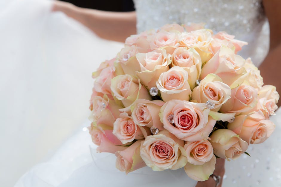 A close-up of a bridal bouquet featuring a dense cluster of pale pink and cream roses, accented with small sparkling wedding jewelry embellishments. The bouquet is held by a person dressed in a white 