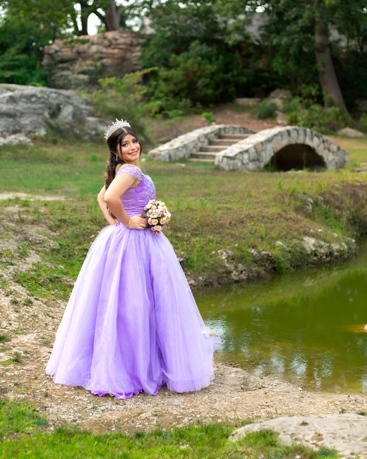 A young woman wearing a purple lavender ball gown with lace detailing on the bodice and short flutter sleeves, standing outdoors near a small pond in a park setting. She is holding a bouquet of pale p