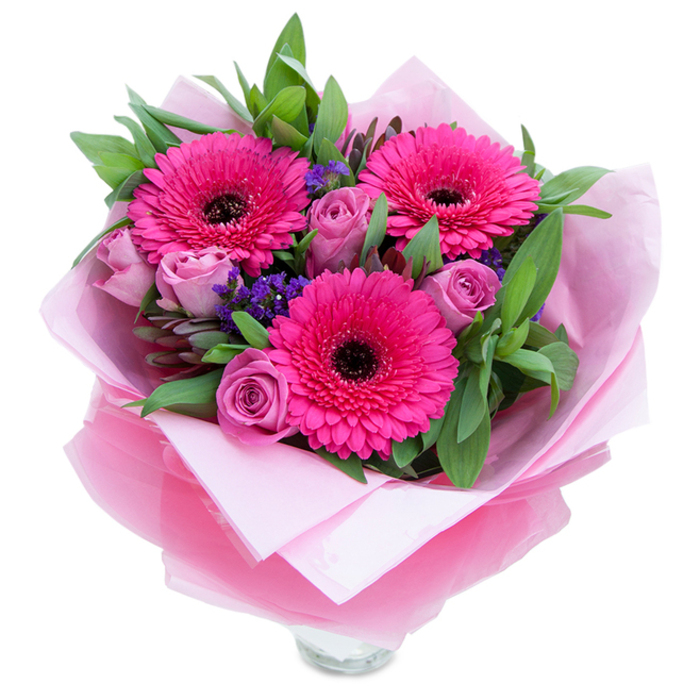 I picture this bouquet as a practiced composition from an experienced local hand: three intense fuchsia Gerbera daisies burst visually forward, their layered petals forming a bright, confident trio with dark central discs that give the design immediate rhythm. Nestled close are several lavender-pink roses in a slightly dusty tone, their petals tightly wound and velvety to the eye, suggesting a soft, classic rose scent that will be noticed when the bouquet is unwrapped. Sprays of small purple statice provide airy punctuation and a contrasting cool tone that makes the pinks sing, while broad, verdant foliage-likely salal or eucalyptus-builds volume and a dewy green backdrop. The floristry is careful: the blooms rest on an inner sheet of pale pink matte tissue, and an outer layer of sheer fuchsia-pink cellophane, slightly crinkled, wraps the stems in a shimmering finish. The clear glass vase rim visible at the bottom implies immediate display and a freshness that arrives ready to stand on a hall table or bedside cabinet. Under soft natural light from the shop window on Friern Barnet Lane, textures and hues are gentle but vivid, petal edges catching faint highlights. As a piece intended for birthdays, anniversaries, or to say 'thank you' to someone special in the local area-North Finchley and beyond-this bouquet balances playfulness with grace. The overall effect is intimate and uplifting: colour, scent, and hand-crafted wrapping designed to comfort and delight the recipient.